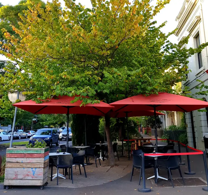 Red umbrellas and footpath tables under a big leafy street tree outside Panette Eatery