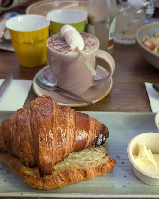 A croissant with butter and jam, hot chocolate behind